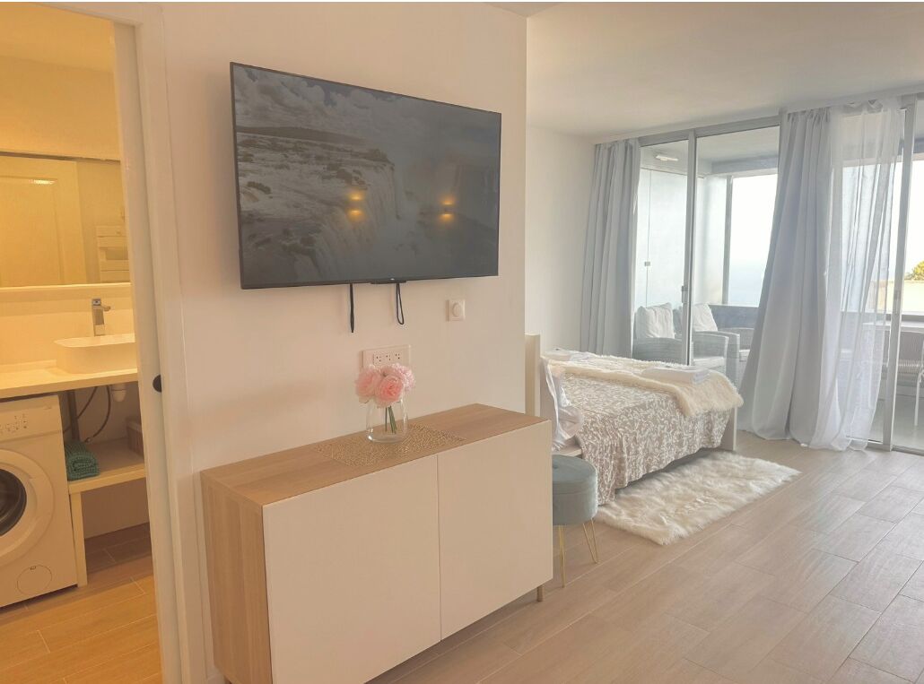 A modern hotel room with a flat-screen TV mounted on the wall, a wooden console table with a vase of flowers, a bed with a white blanket, and a balcony with a view.