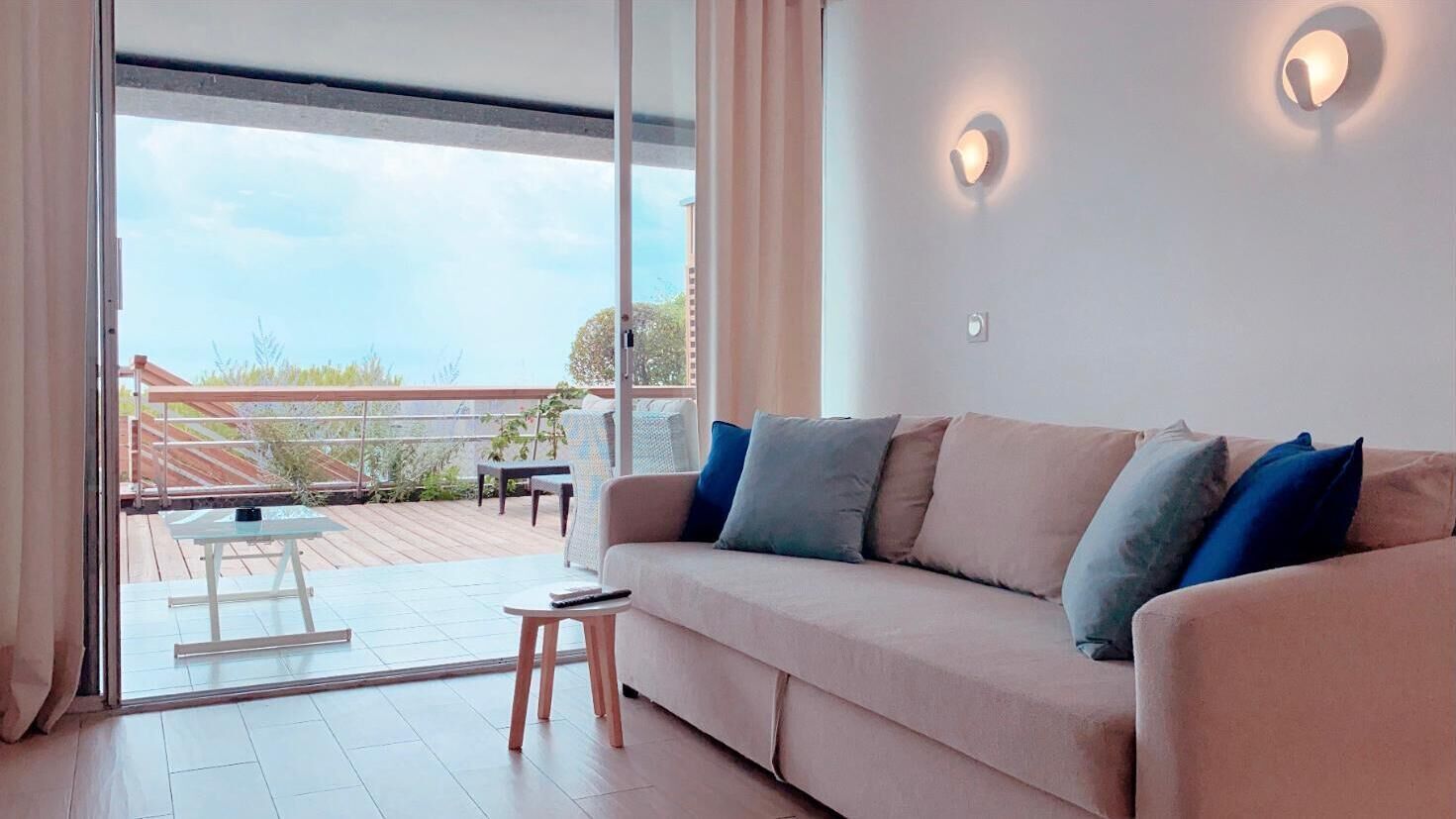 Ein modernes Wohnzimmer mit einem beigen Sofa, zwei Kissen, einem Glastisch und einem kleinen Beistelltisch. Eine Glastür führt auf eine Terrasse mit Tisch und Stühlen.