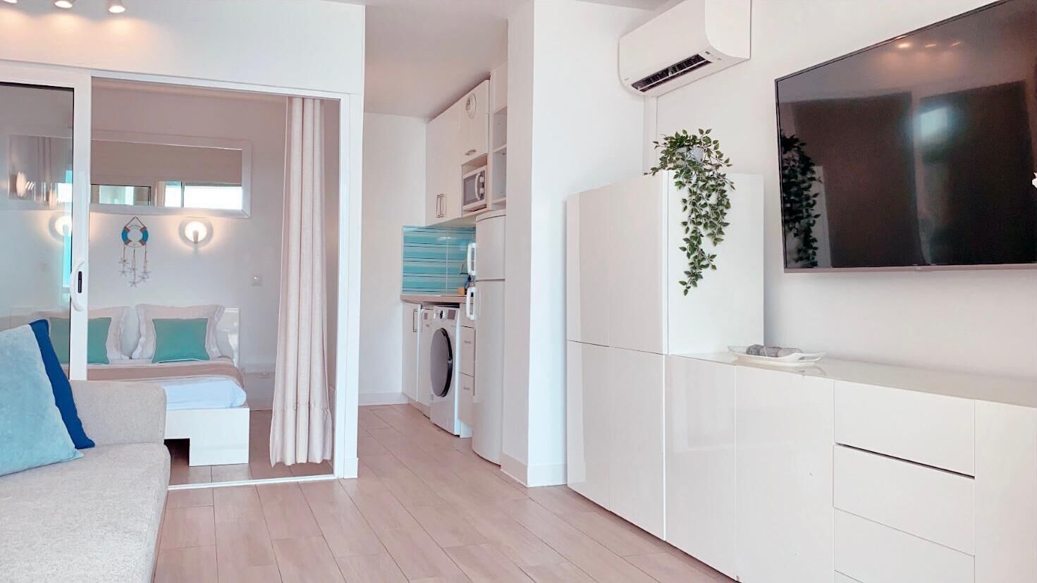 A modern apartment with a living area, a kitchen with a built-in washer and dryer, and a large TV mounted on the wall.