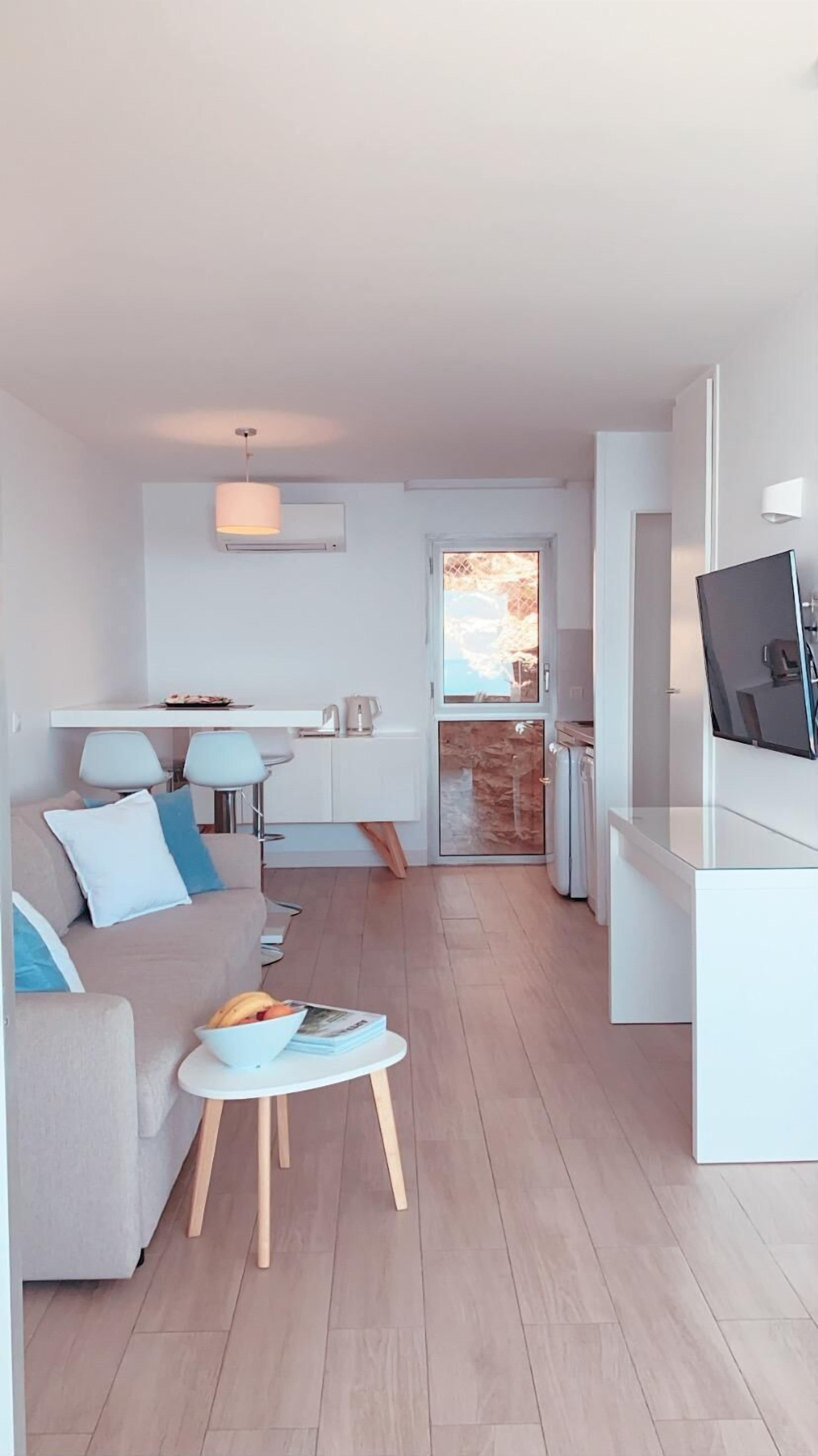 Un salon moderne avec un canapé, une table basse et un téléviseur. Il y a un coin cuisine avec une table à manger et des chaises. La pièce est dotée d’une grande fenêtre et d’une porte donnant sur un balcon.