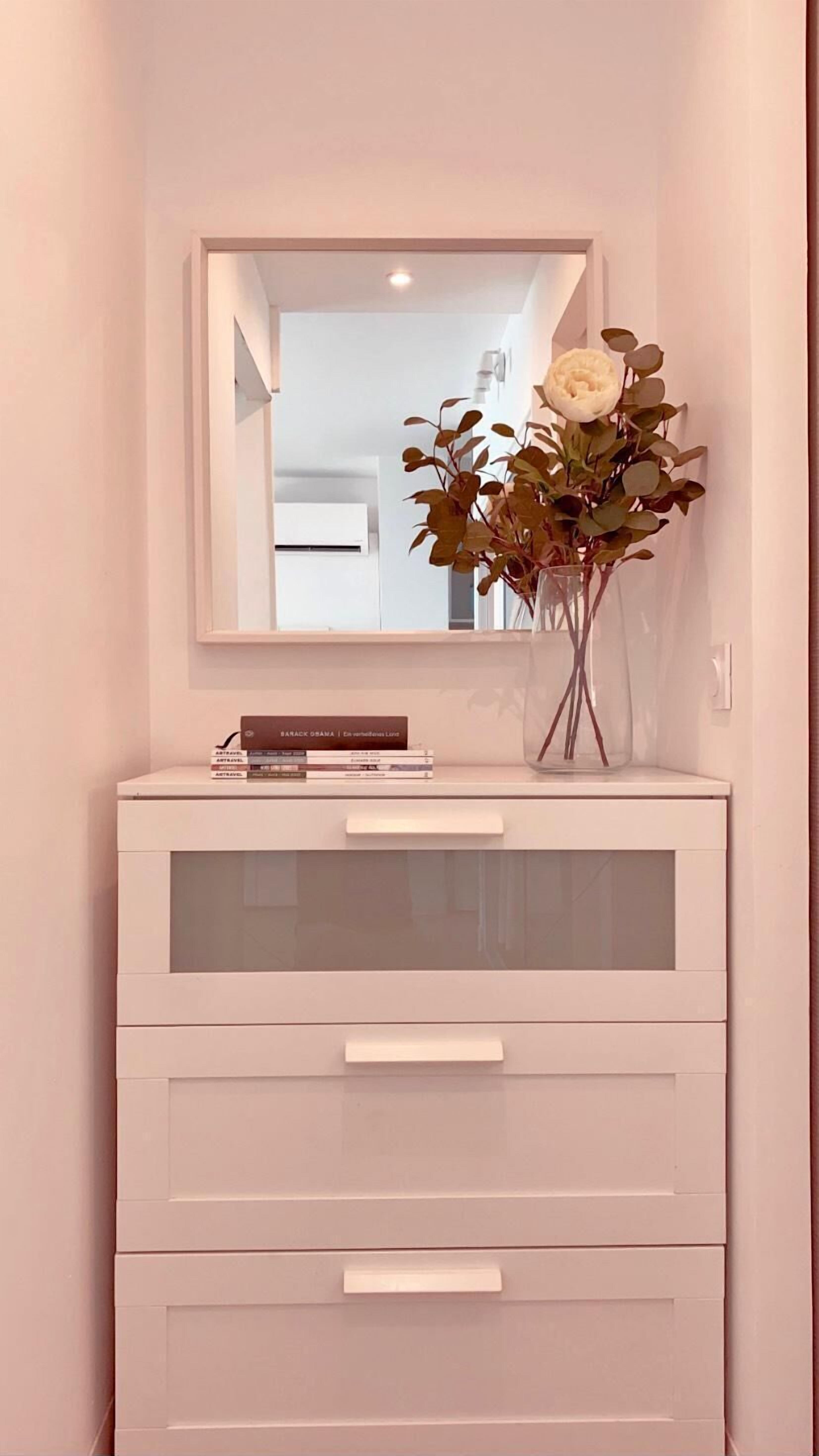 A white vanity with a mirror, a vase with dried flowers, and a stack of books.