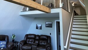 Un'ampia zona giorno con soffitto alto, travi a vista in legno, una ventola a soffitto, una scala che conduce a un livello superiore e un divano in pelle marrone.