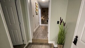 Un corridoio con pavimento in legno, una piccola scala, una pianta in vaso e una porta con l'interruttore della luce.