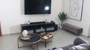 Een moderne woonkamer met een flatscreen televisie aan de muur, een grijze bank, een ronde salontafel met een schaal lekkernijen, en een minimalistisch kunstwerk.