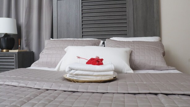 Een opgemaakt bed met een grijze gewatteerde sprei, twee kussens en een stapel witte handdoeken met een rode bloem op een dienblad.