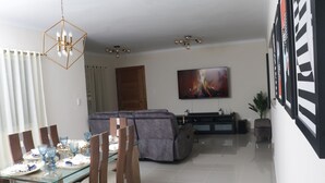 Een moderne woonkamer met een glazen eettafel, houten stoelen, een grijze bank en een flatscreen televisie aan de muur.
