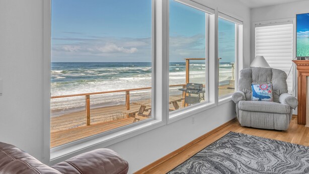 A room with large windows overlooking a beach, a gray armchair, a wooden table, and a patterned rug.