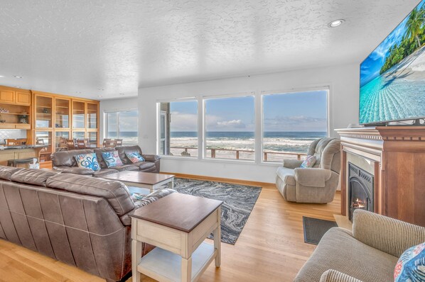 A spacious living room with a large window overlooking the beach, a fireplace, and a kitchen area with wooden cabinets.