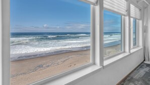 A room with a large window overlooking a beach with waves and a clear sky.