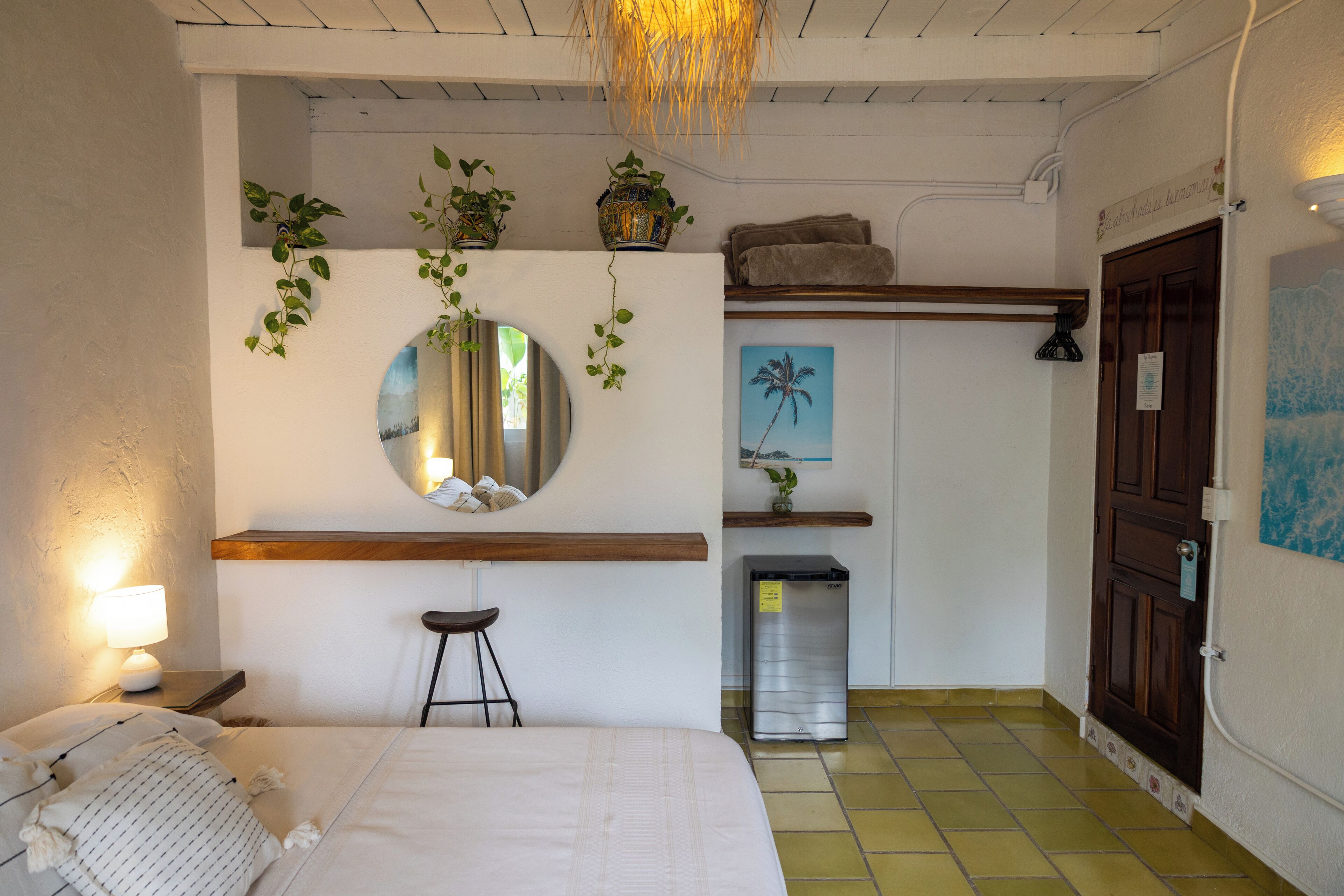 Un dormitorio con cama, mesita de noche, lámpara, espejo, plantas en macetas y un cubo de basura.
