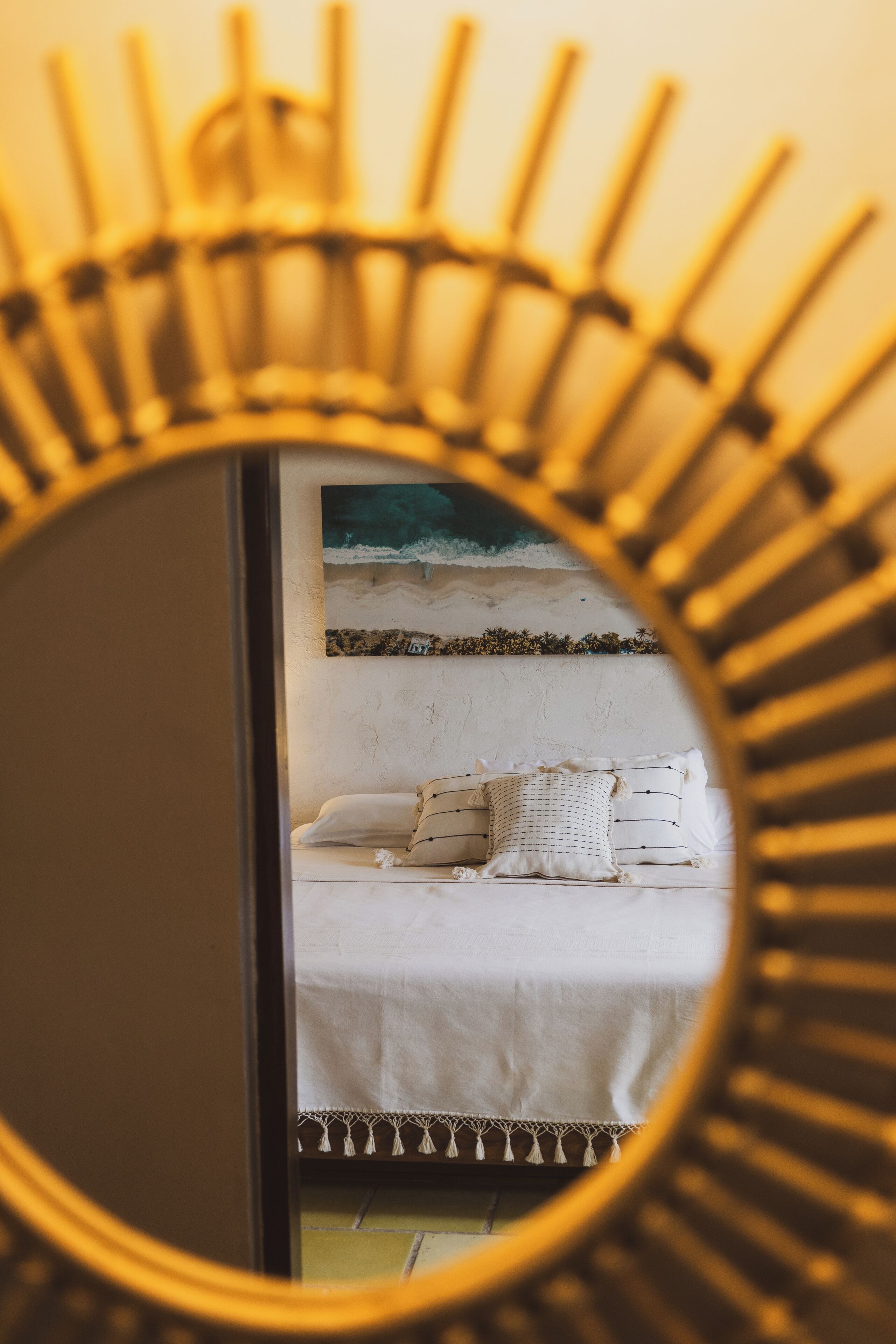 Een bed met wit beddengoed en een kussen met een patroon, een muur met een schilderij en een spiegel met een zonachtige omlijsting.