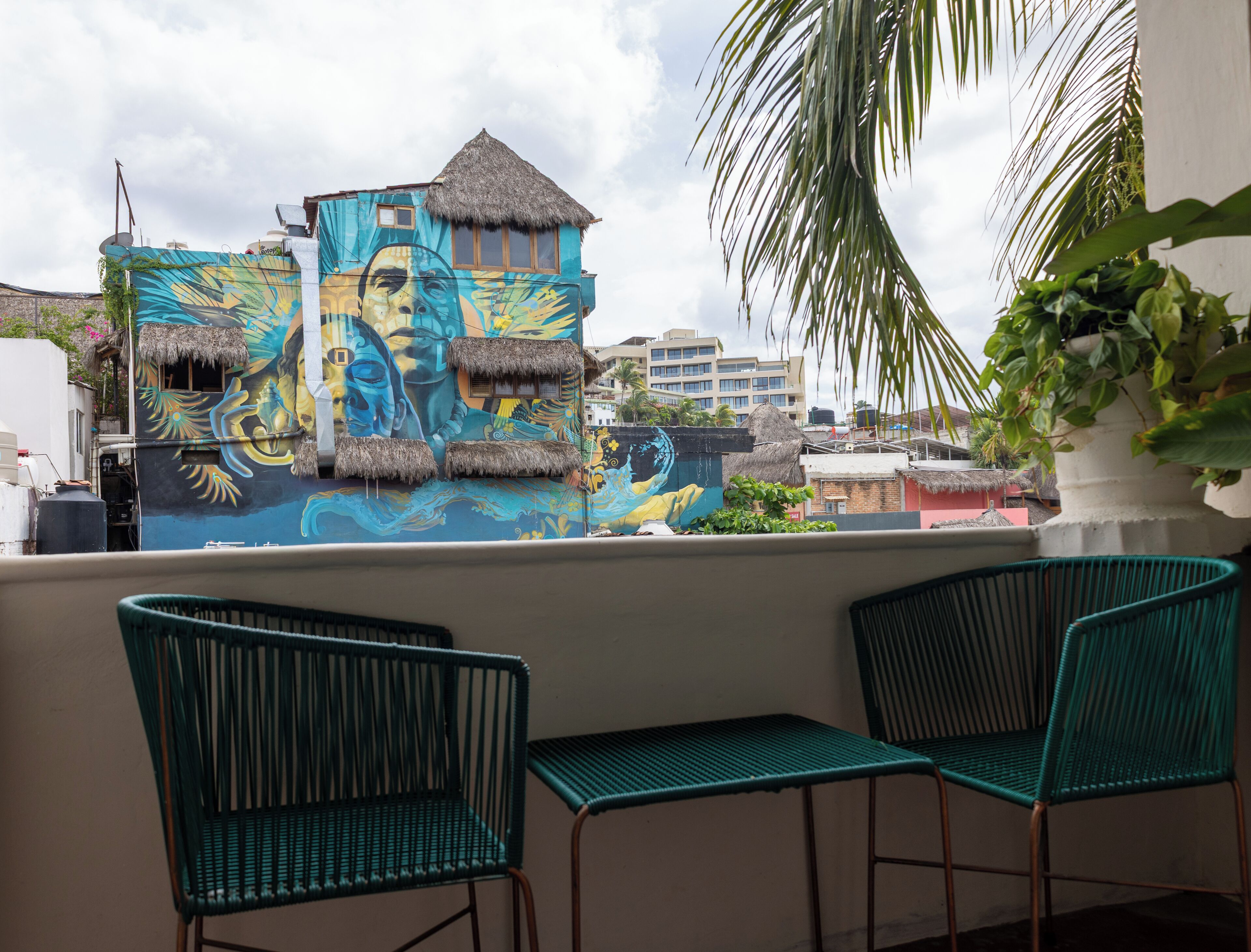 A balcony with two green chairs, a mural with tropical and oceanic designs, and a view of buildings in the background.