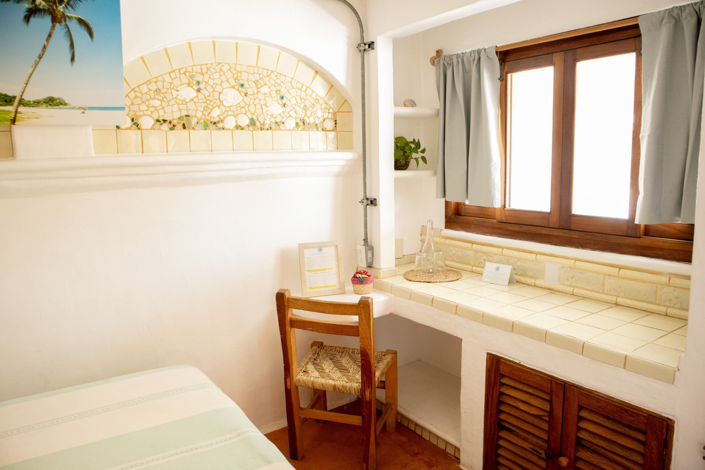 A bathroom with a tiled vanity, a wooden chair, a window with curtains, and a decorative mosaic wall.