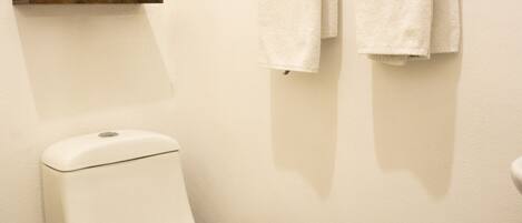 Een schone, witte badkamer met een toilet, een klein plankje erboven en twee handdoeken aan een rek.