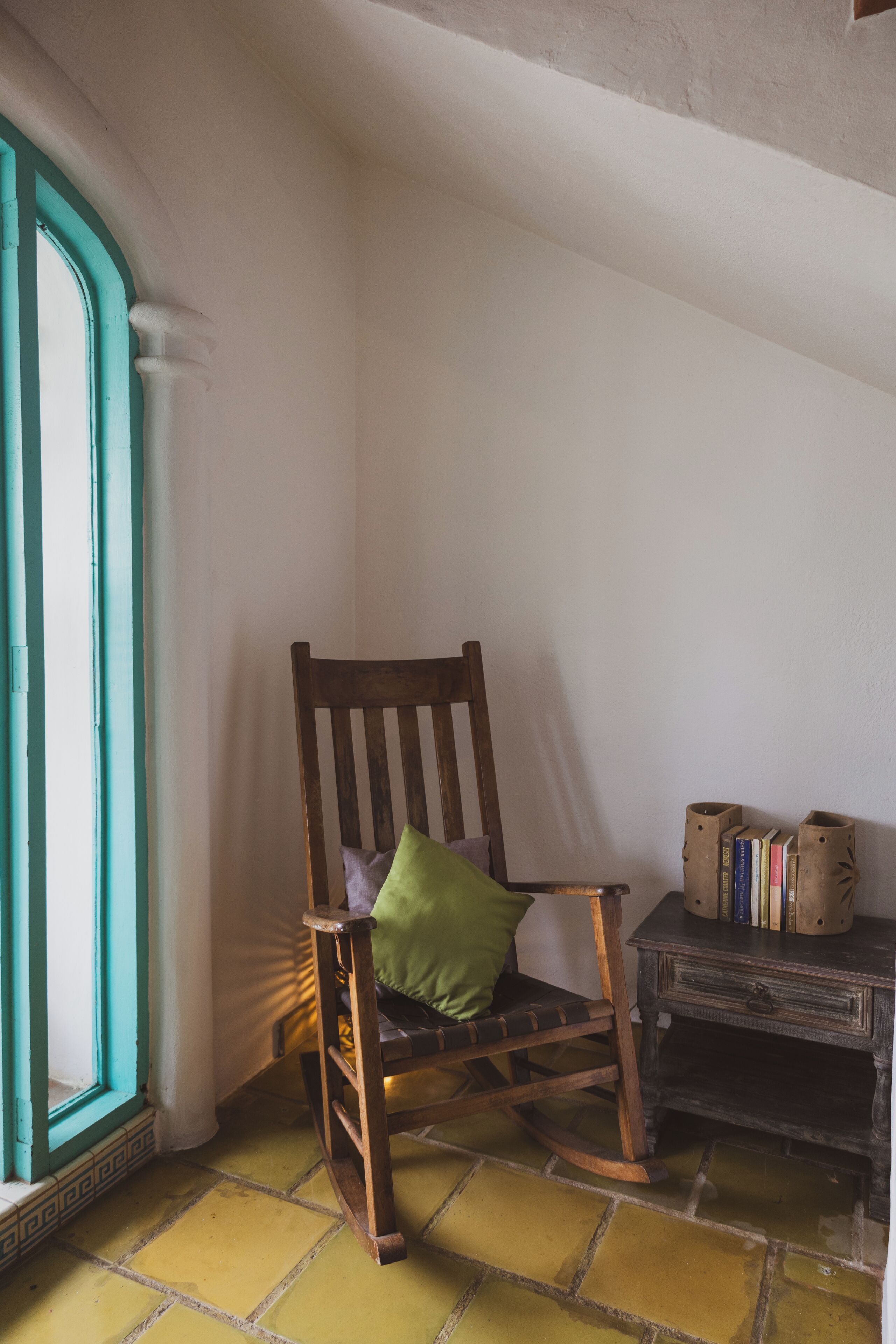 A corner room with a wooden chair, a small table with books, and a window with a teal frame.