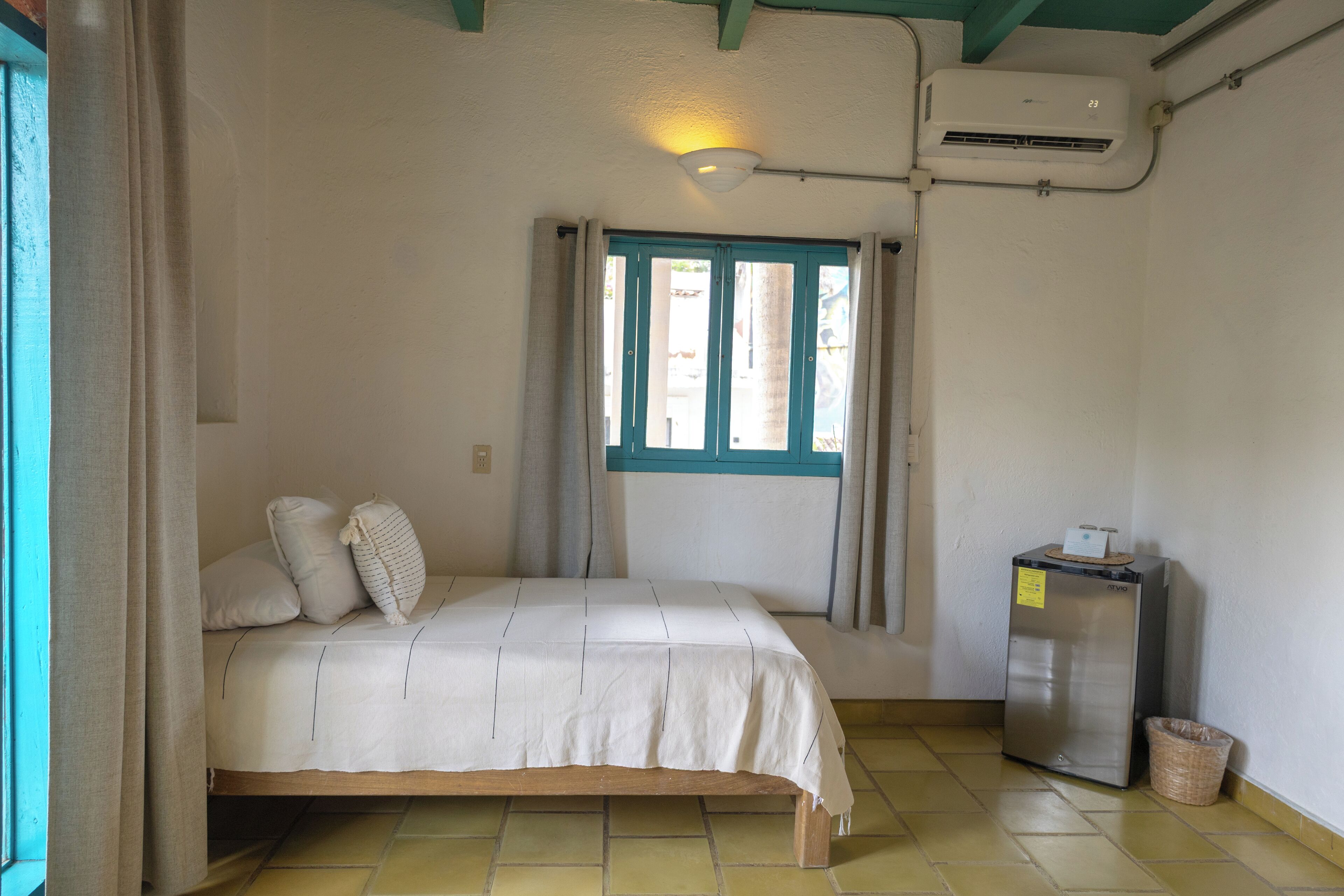 A room with a bed, a window with curtains, a small refrigerator, and a trash bin.