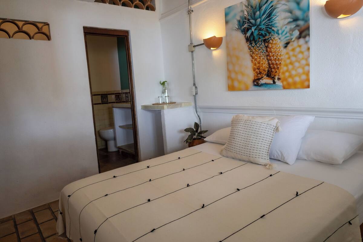 A bedroom with a bed, a nightstand, a potted plant, and a wall-mounted light fixture.