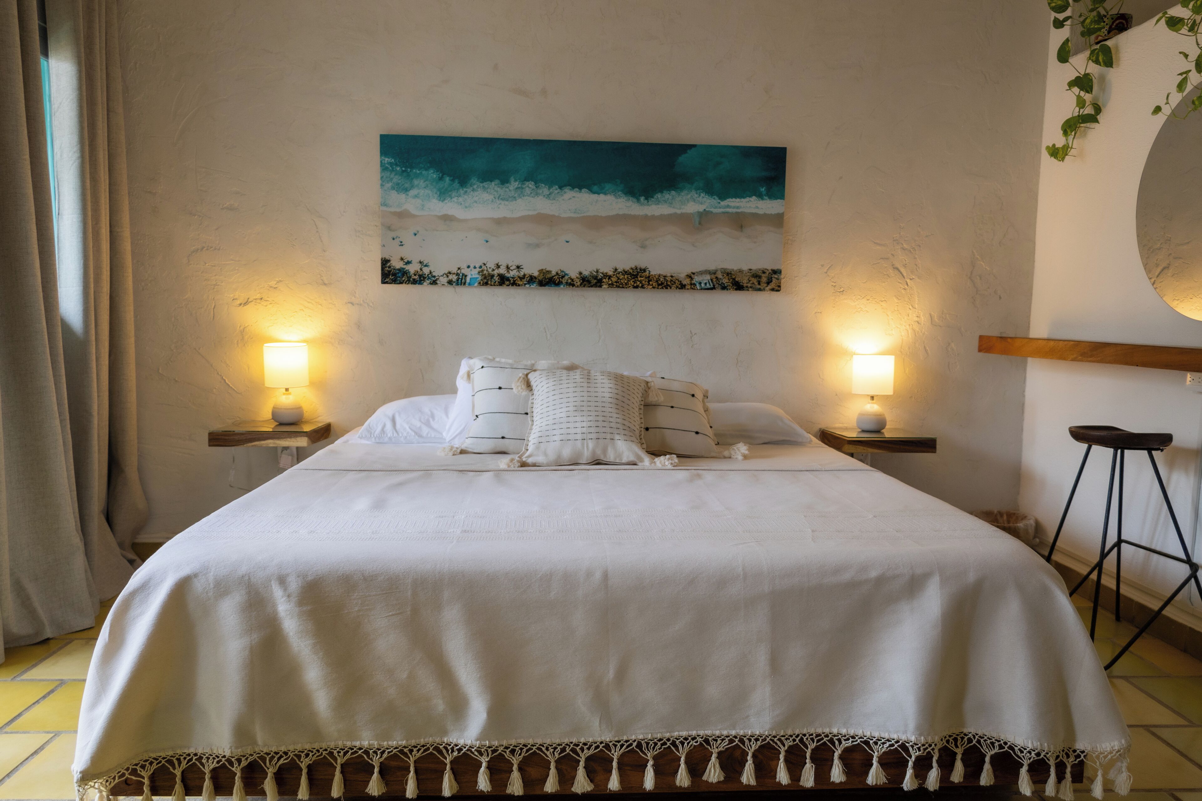 A neatly made bed with a white bedspread and pillows, two bedside lamps, a painting of a beach scene, and a round mirror on the wall.
