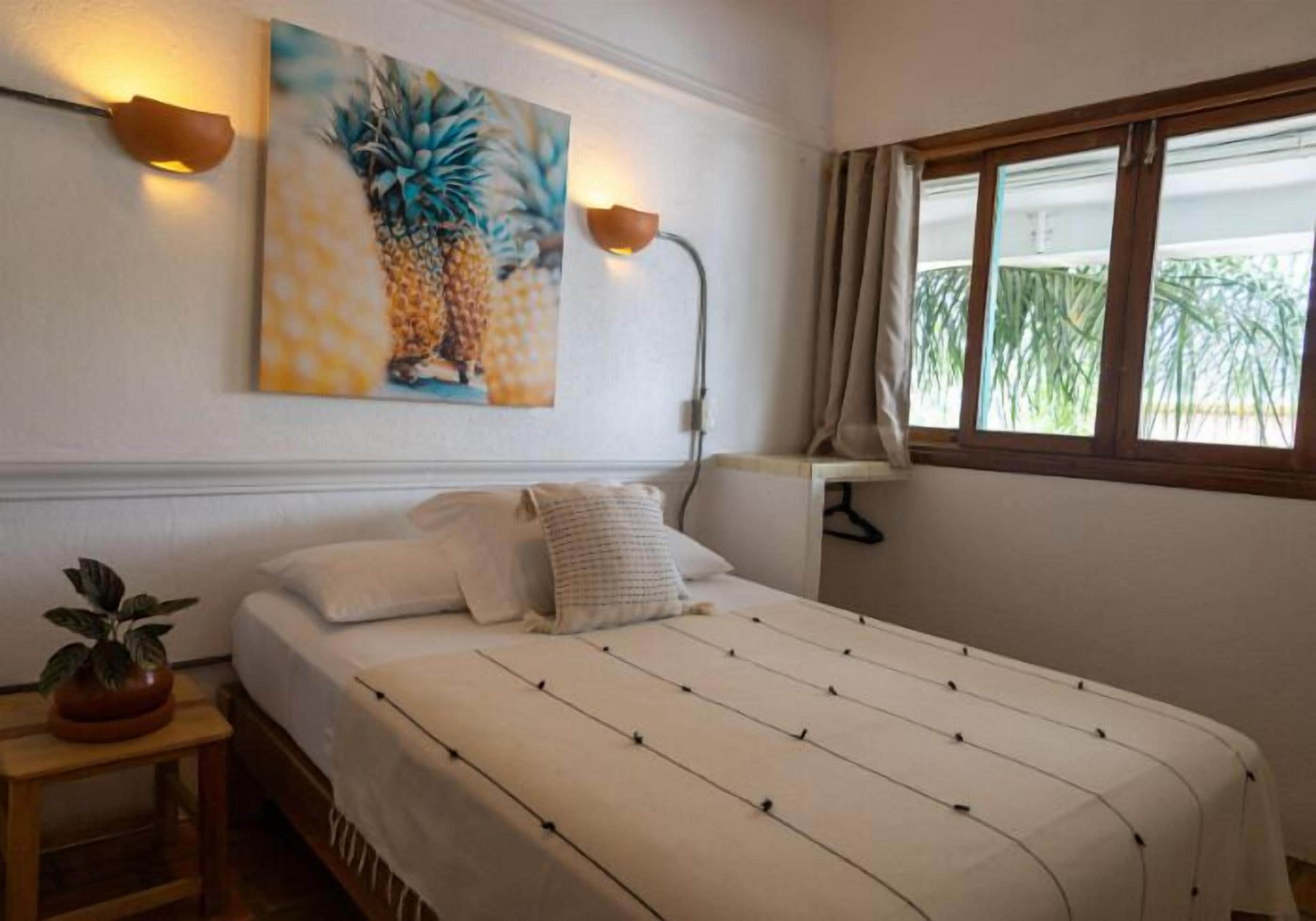 Un dormitorio con una cama, un cuadro de piñas, una lámpara de noche, una ventana con cortinas y una planta en una mesita de noche.