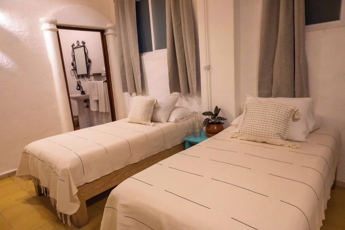 A bedroom with two beds, a small table with a plant, and a bathroom visible through an archway.