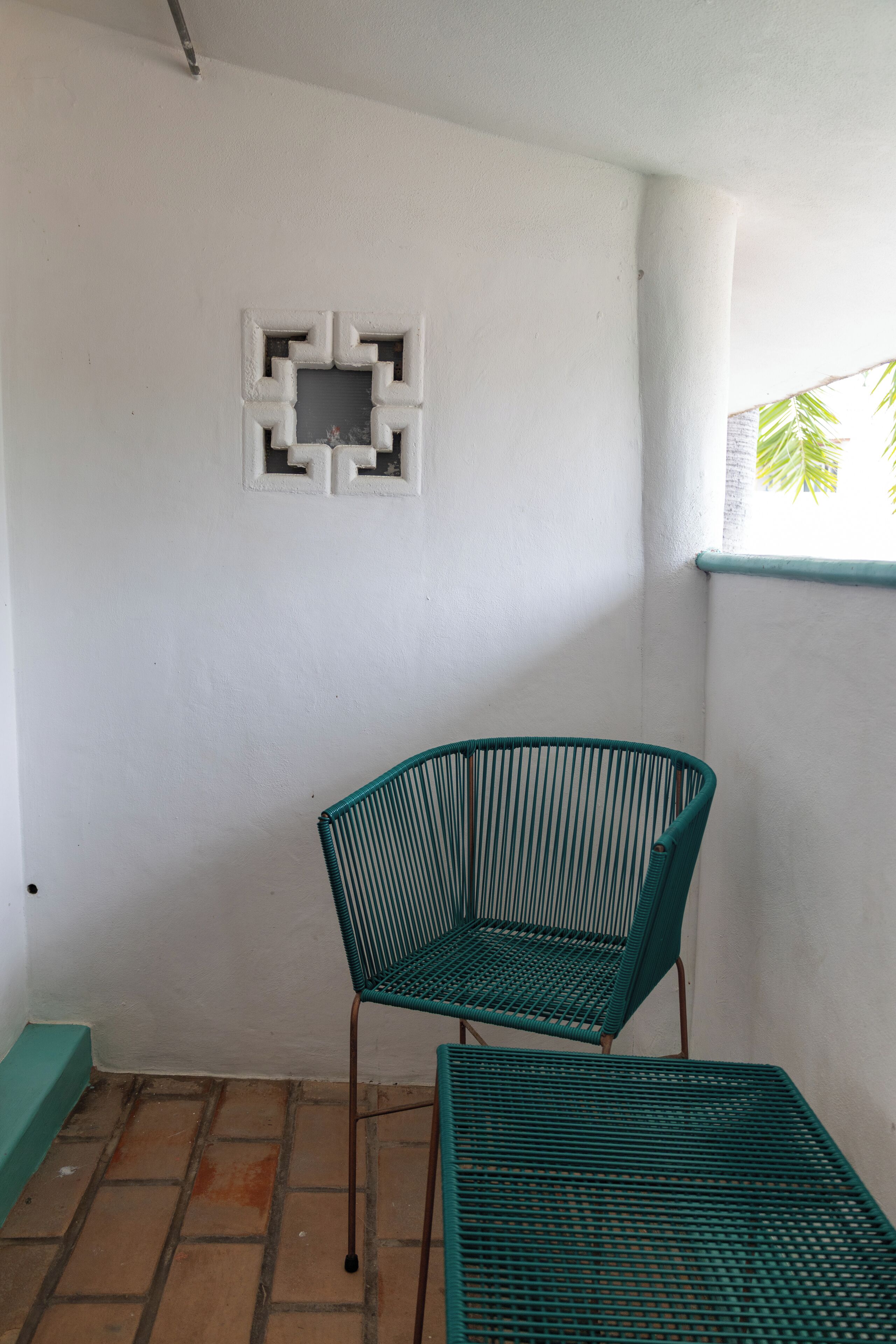 Ett rum med en grön metallstol, ett litet bord, en vit vägg med ett geometriskt mönster och ett fönster med utsikt över lövverket.