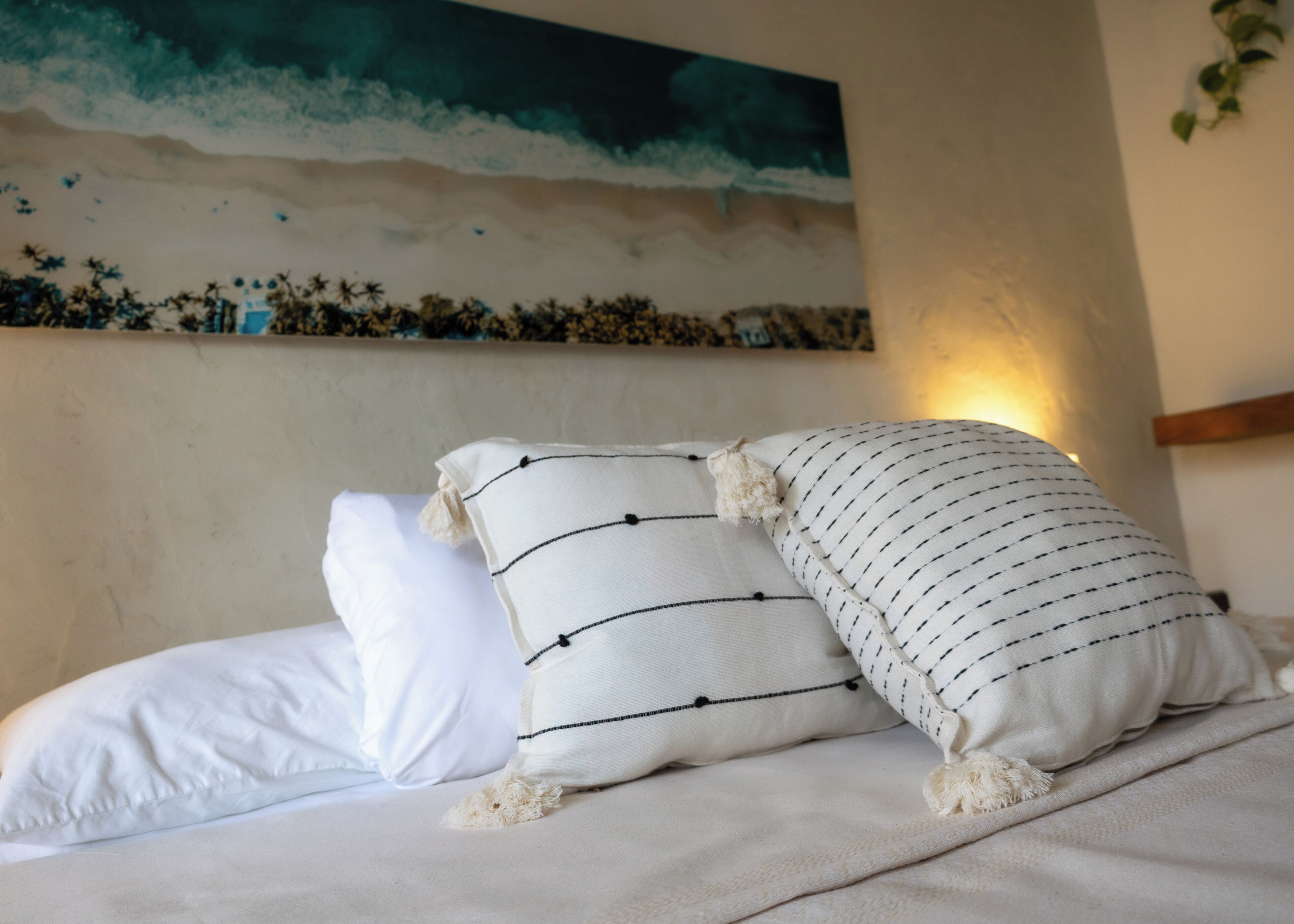 A bed with white linens and striped pillows, a painting of a beach scene on the wall, and a plant on the right.