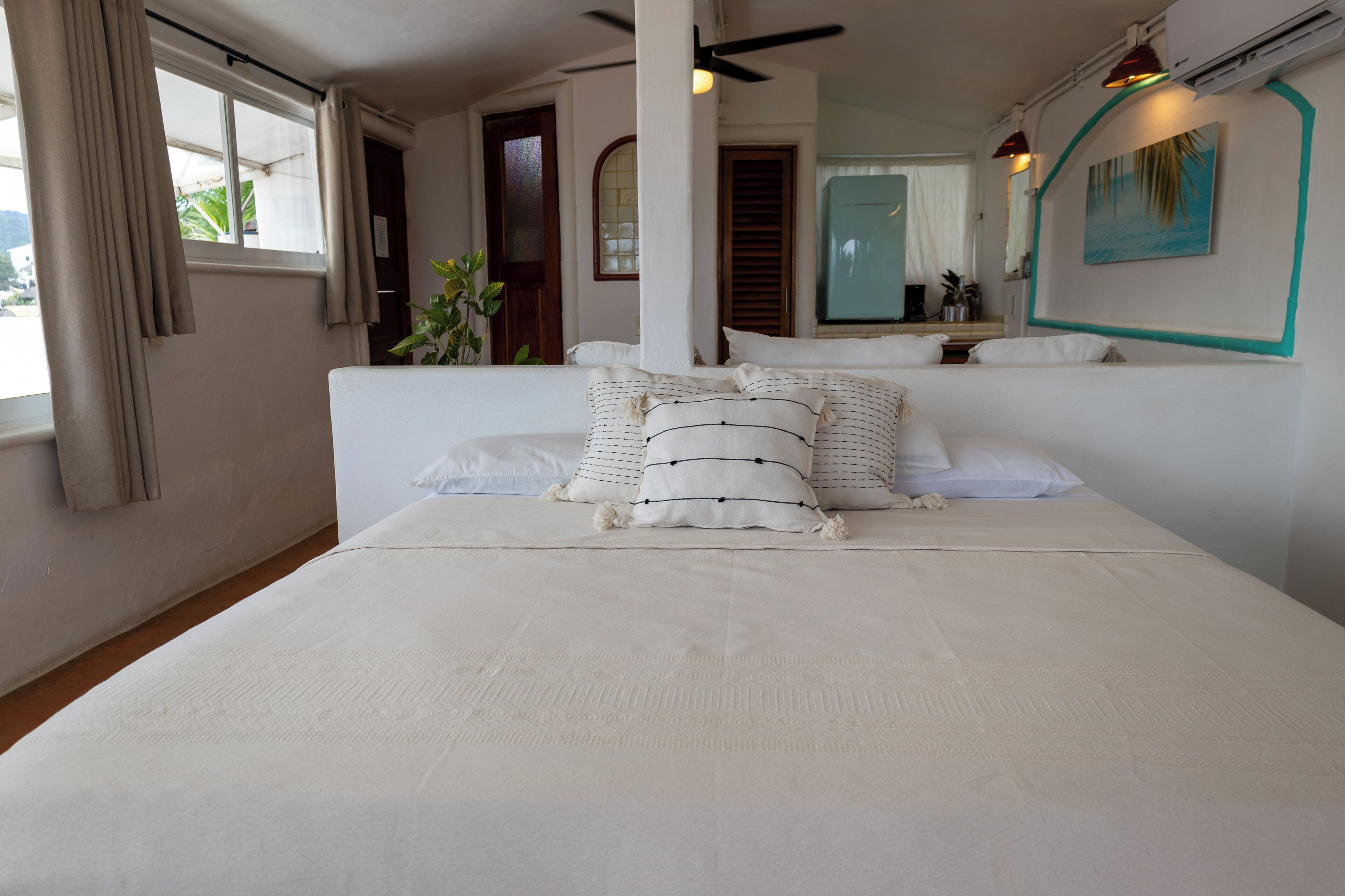 A neatly made bed with white bedding and striped pillows, a window with curtains, and a painting on the wall.