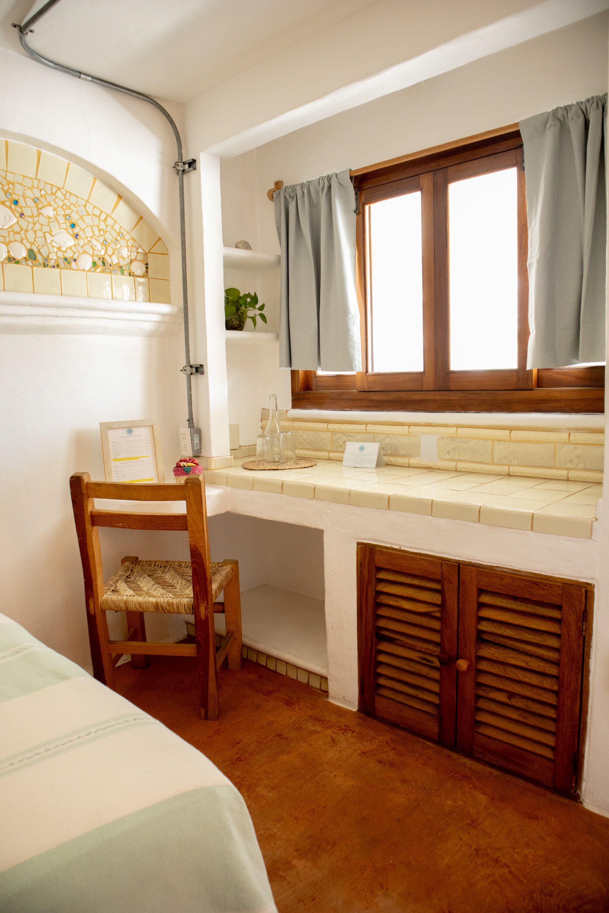 Una habitación con una silla de madera, una mesita pequeña, una ventana con cortinas, un rebozo de azulejos y una puerta de madera.
