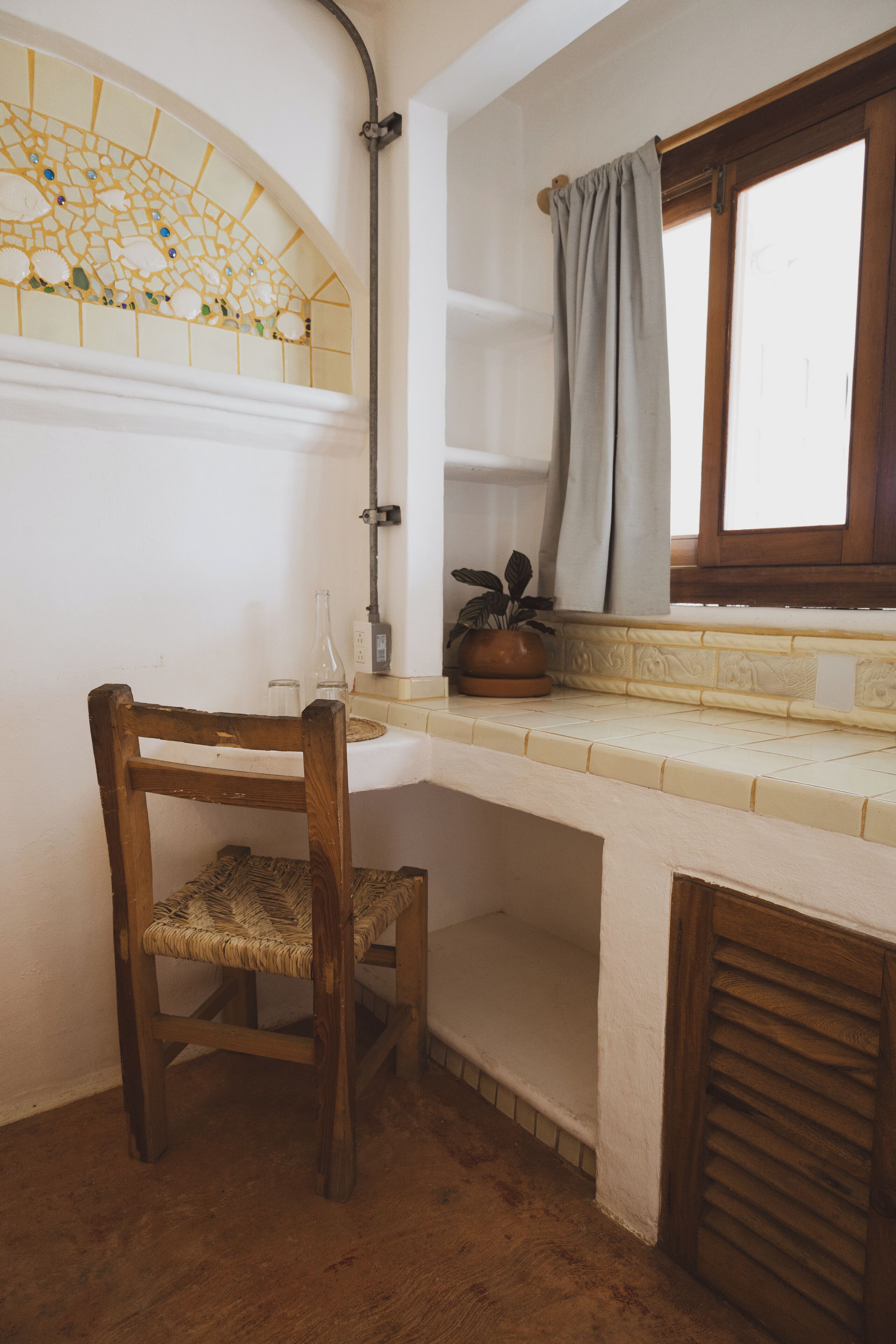 A corner room with a wooden chair, a small table, a potted plant, a window with a curtain, and a tiled backsplash.