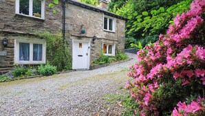 Une petite maison en pierre avec une allée en gravier, une porte blanche et un petit jardin agrémenté de fleurs roses.