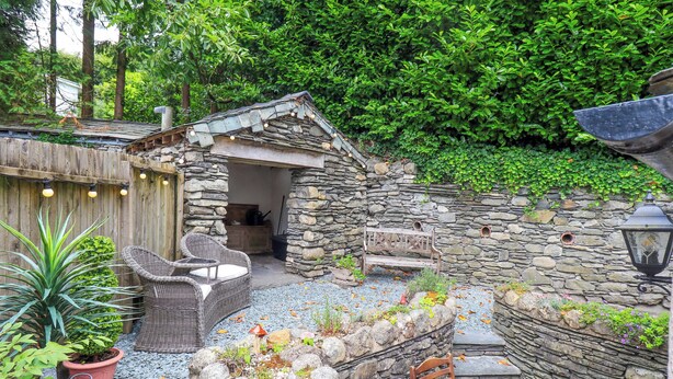 Un espace extérieur aménagé avec des murs en pierre, comprenant un banc et des chaises, et entouré de plantes en pot et d’un mur de pierre.