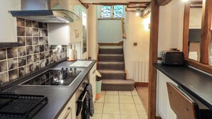 Une cuisine compacte équipée d’une cuisinière à gaz, d’un four encastré et d’un petit plan de travail. Un escalier mène à un niveau supérieur, et une fenêtre offre une vue sur la verdure environnante.