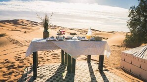 Eine Wüstenlandschaft mit einem Tisch, der für eine Mahlzeit gedeckt ist, umgeben von Sanddünen und einem klaren Himmel.