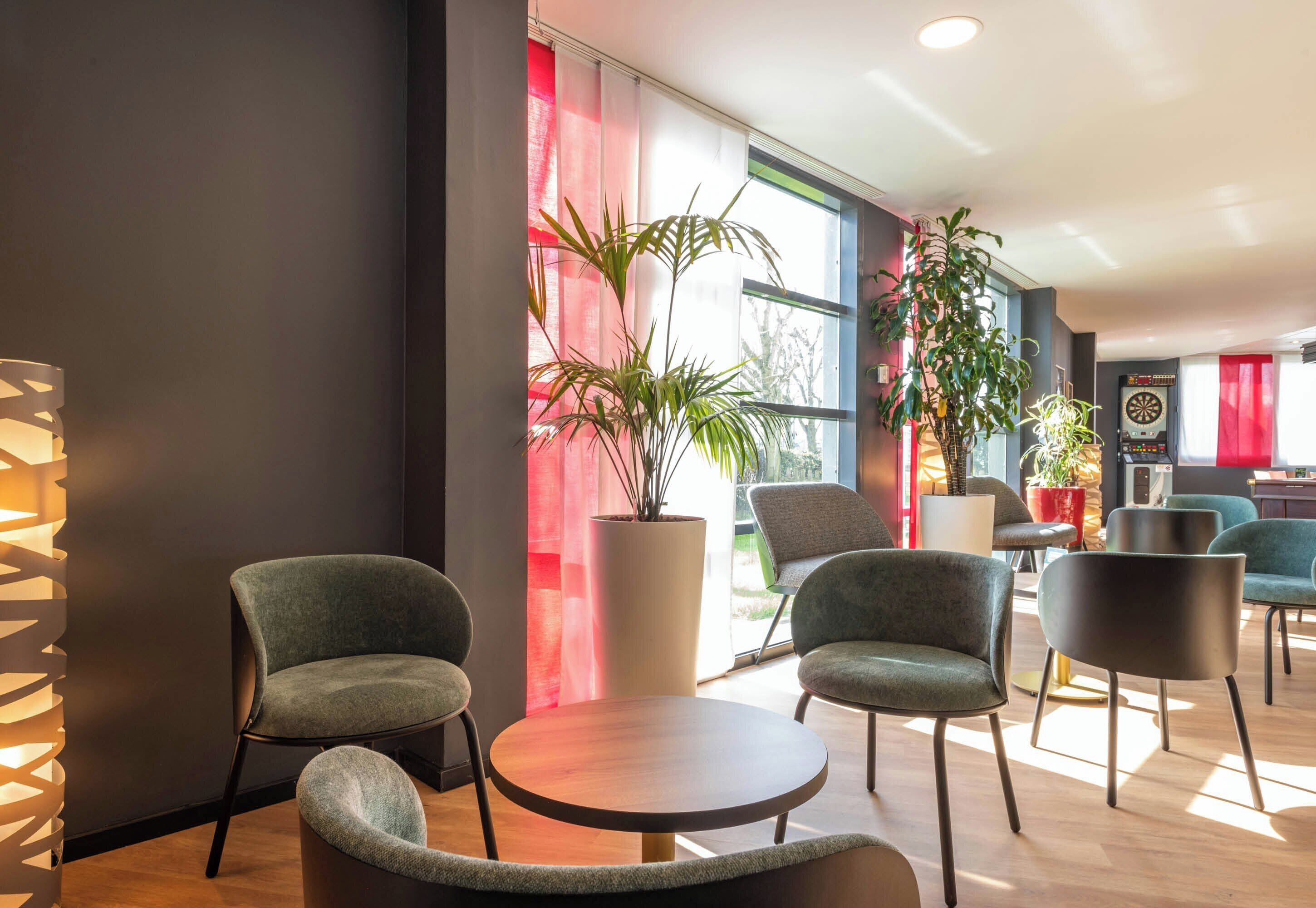 A modern lounge area with round tables and chairs, large windows with red curtains, and potted plants.