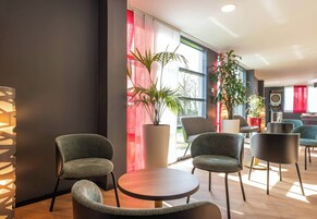 Un área de descanso moderna con mesas y sillas redondas, ventanas amplias con cortinas rojas y plantas en macetas.