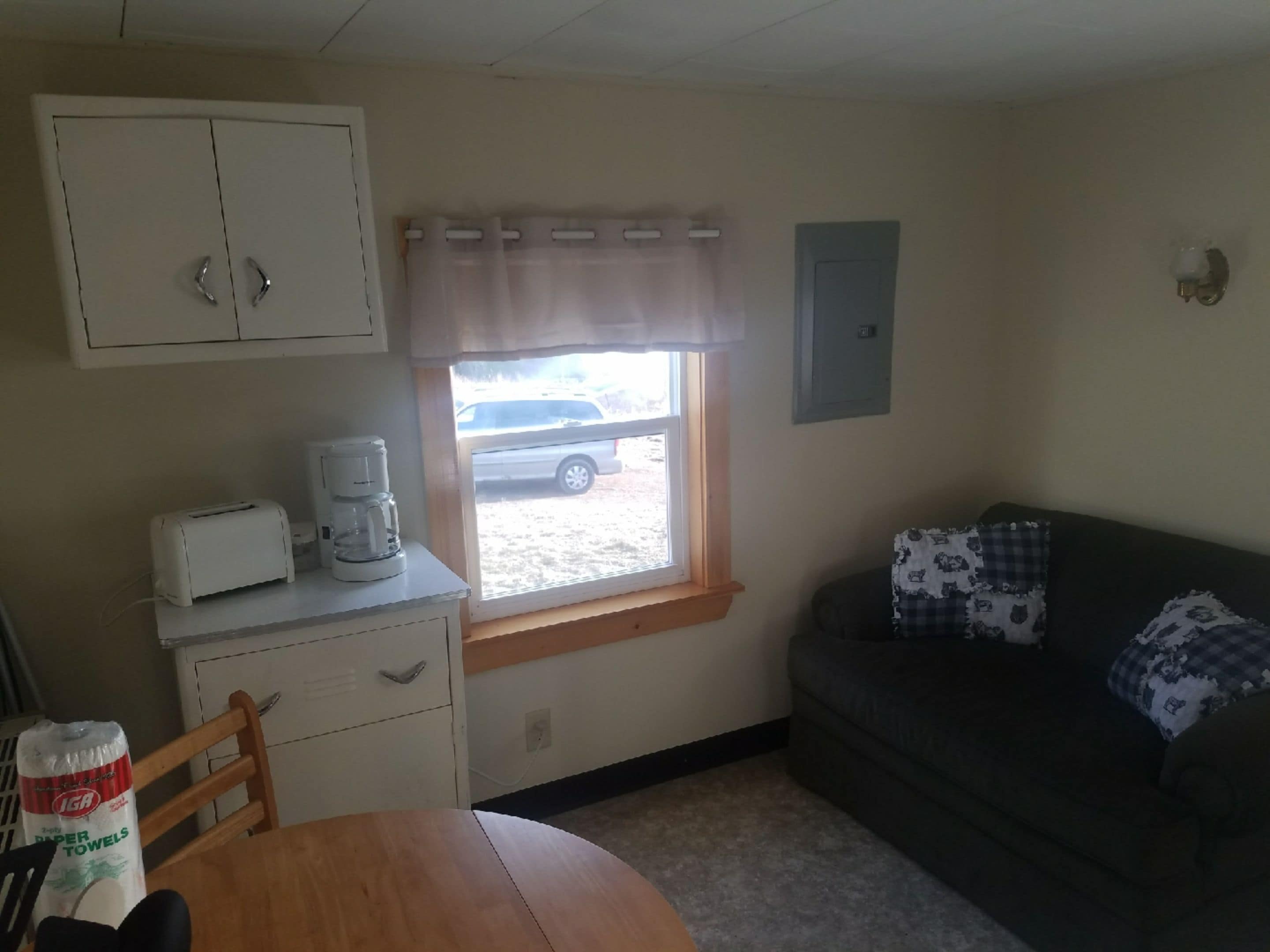 A small, compact room with a kitchenette area including a toaster and coffee maker, a window with a view of a parked car outside, a dark-colored sofa with patterned cushions, and a wooden dining table.