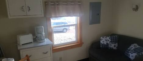 A small, compact room with a kitchenette area including a toaster and coffee maker, a window with a view of a parked car outside, a dark-colored sofa with patterned cushions, and a wooden dining table.