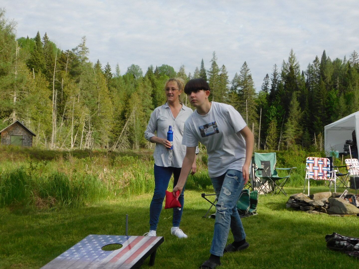 Two people standing near a small wooden structure in a grassy area with trees in the background.