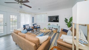 A spacious living room with a large brown sofa, two blue armchairs, a wooden coffee table, and a television mounted on the wall.
