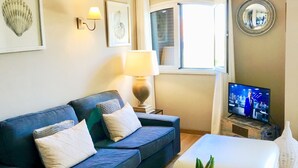 Una sala de estar con un sofá azul, una pequeña mesa redonda con una planta, un televisor sobre un mueble de madera, una ventana con cortinas y obras de arte enmarcadas en la pared.