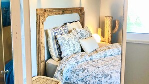 Un dormitorio con cabecera de madera, ropa de cama estampada con flores, dos lámparas de pared, un sillón de mimbre y una ventana con luz natural.