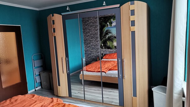 A bedroom with a bed, a wardrobe with a mirror, and a view of a tropical scene through the wardrobe doors. - FeWo - Feeling at home (Wilhelmshaven)
