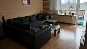 A living room with a black sofa, a glass coffee table, and a radiator. There is a window with curtains and a view of the outside. - FeWo - Feeling at home (Wilhelmshaven)