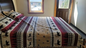A bedroom with a bed featuring a quilt with animal prints, a window with a view of the outdoors, and two bear silhouettes on the wall. - 01) The Bear Cave Cabin (Stewartstown)