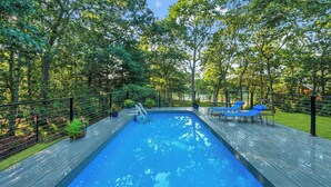 A rectangular swimming pool with a wooden deck, surrounded by trees and a metal fence.