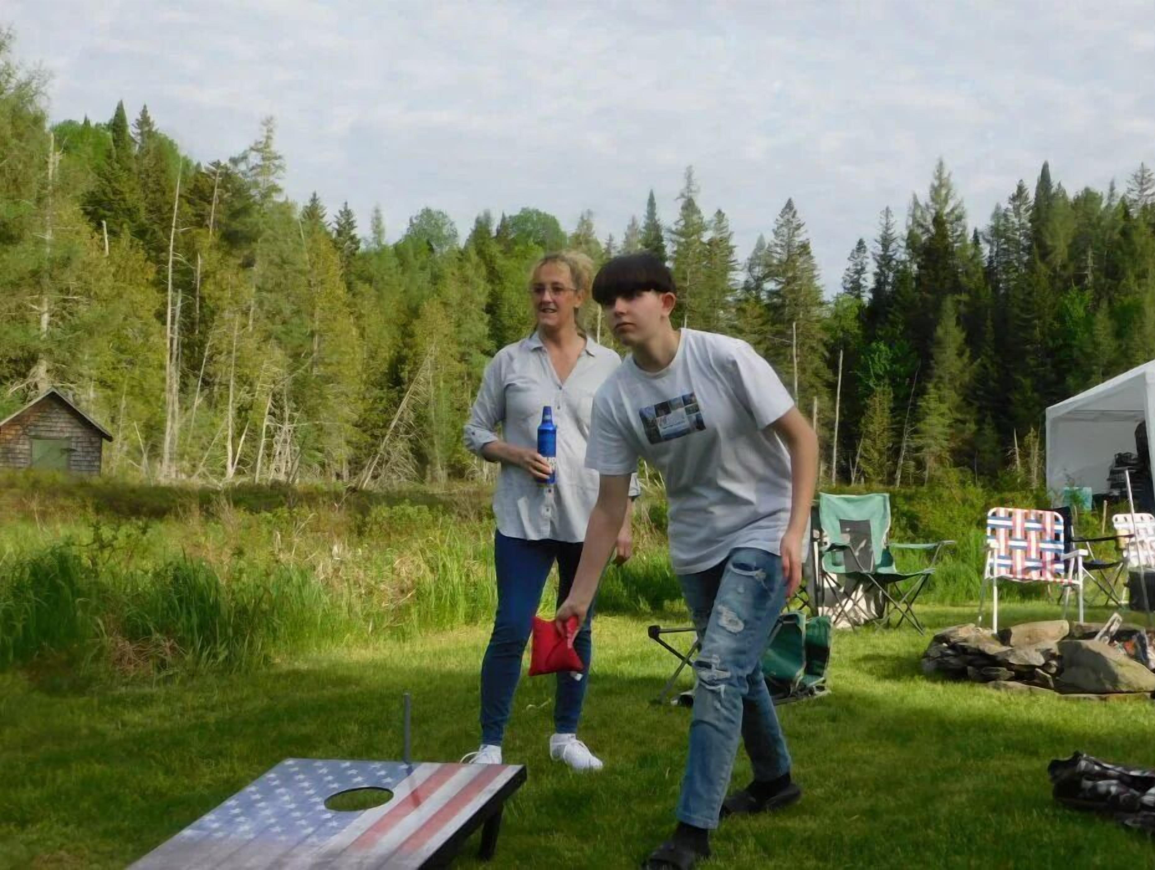 Two people standing in a grassy area with a small American flag on the ground, surrounded by trees and a tent in the background.