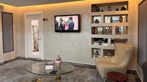 Uma sala de estar moderna com televisão de tela plana, uma mesa de centro de vidro, uma poltrona bege e prateleiras decorativas.