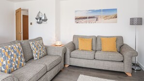 A living room with two grey sofas, one with patterned cushions, a small table with a lamp, and wall art depicting a beach scene. - 31 Beach Road (Porthmadog)