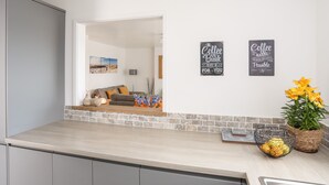 A modern kitchen with a light wood countertop, a grey cabinet, and a view into a living room with a grey sofa and a wall-mounted picture. - 31 Beach Road (Porthmadog)