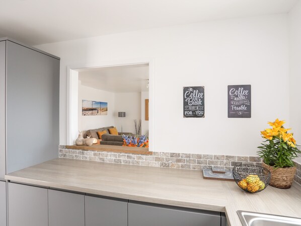 A modern kitchen with a light wood countertop, a grey cabinet, and a view into a living room with a grey sofa and a wall-mounted picture.